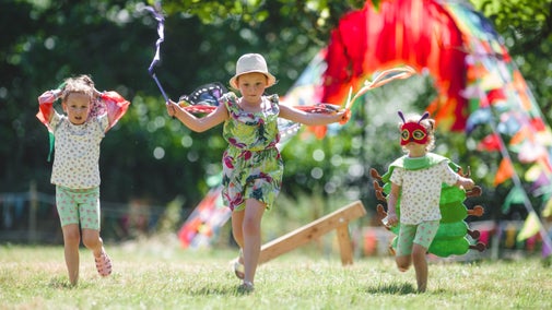 Three young children dress in insect costumes running through a grassy field. In the background their is a rainbow archway made from ribbons.